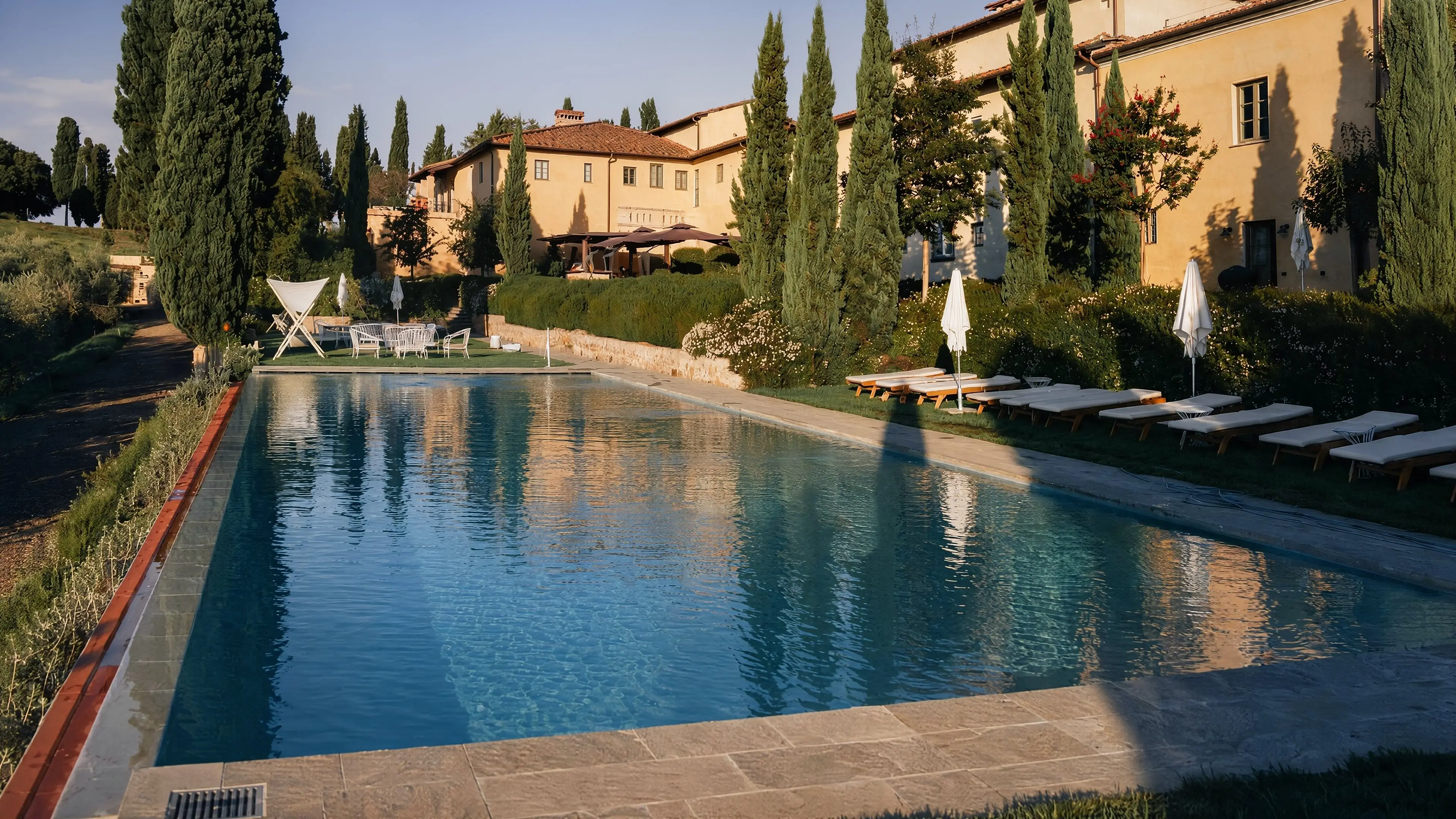 La piscina lunga del podere alla luce bassa del pomeriggio, coi cipressi e il casolare in pietra chiara sul fondo
